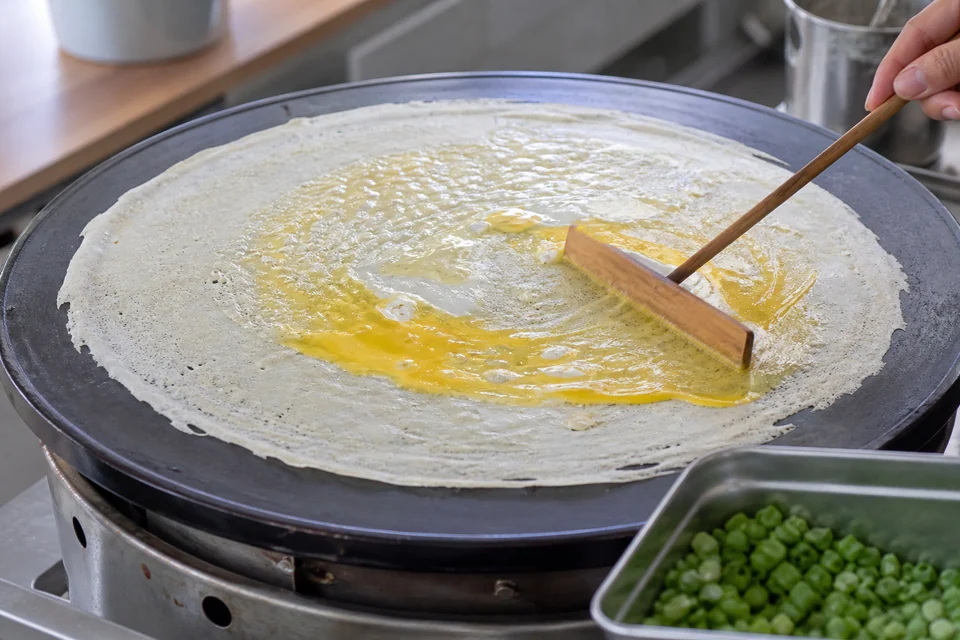 Thin jianbing-style batter being spread on a hot round griddle before egg and scallions are added.