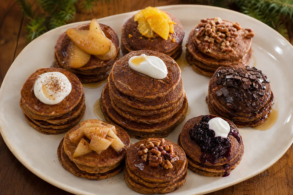 Ten small stacks of finished gingerbread pancakes with pear, orange, pecans, chocolate, apple, berries, and yogurt