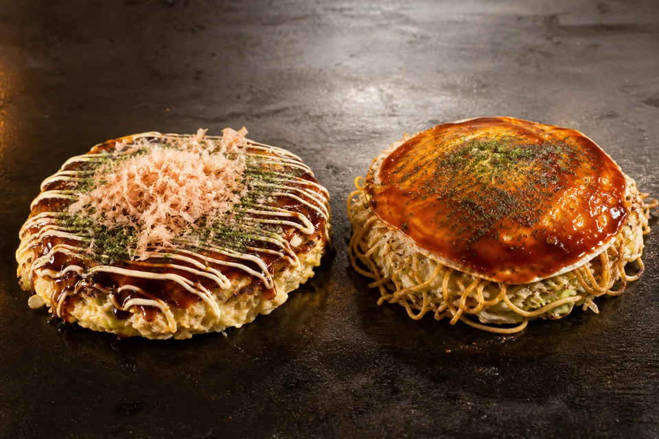 Two finished okonomiyaki on a griddle: one mixed cabbage round and one visibly layered noodle-filled Hiroshima-style round.