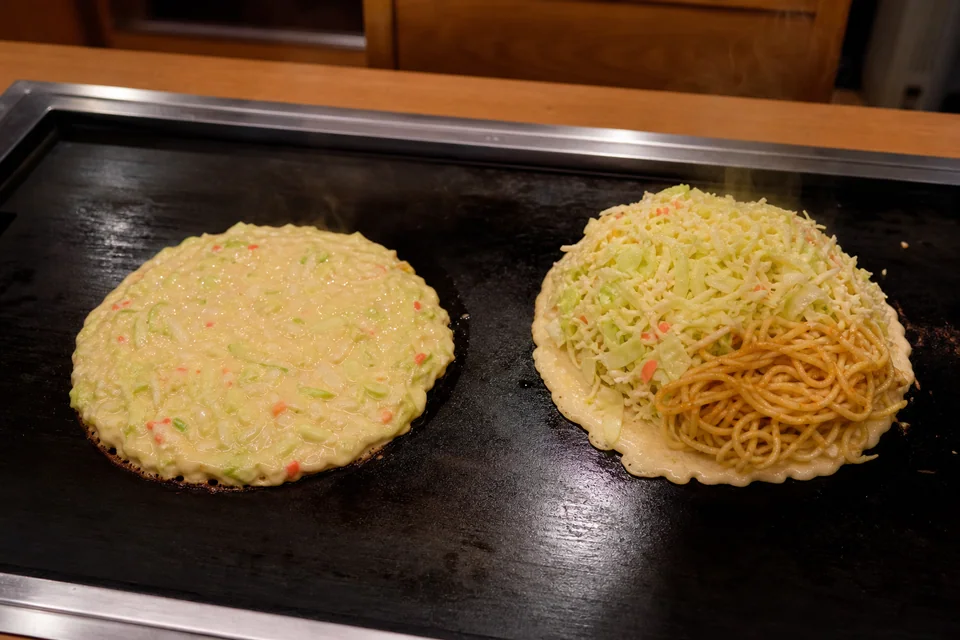 Mixed cabbage batter and a separate thin base with cabbage and noodles cooking side by side to compare okonomiyaki construction.