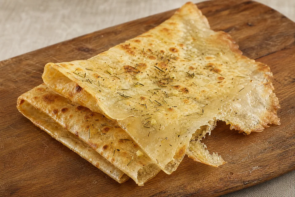 Paper-thin folded borlengo with a lacy torn edge and light garlic-rosemary seasoning on a rustic wooden board.