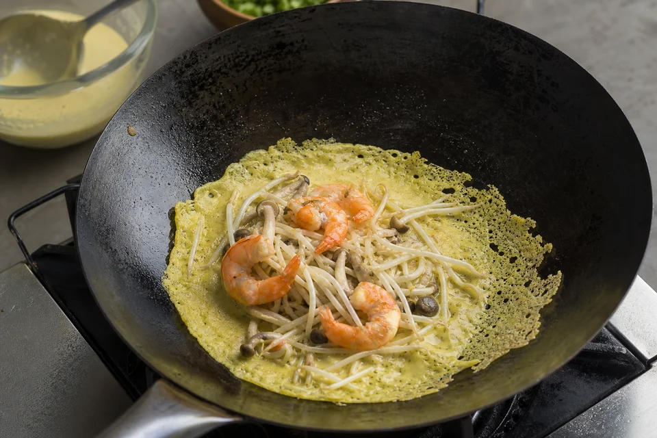 Thin turmeric bánh xèo batter crisping around shrimp, mushrooms, and bean sprouts in a seasoned pan.