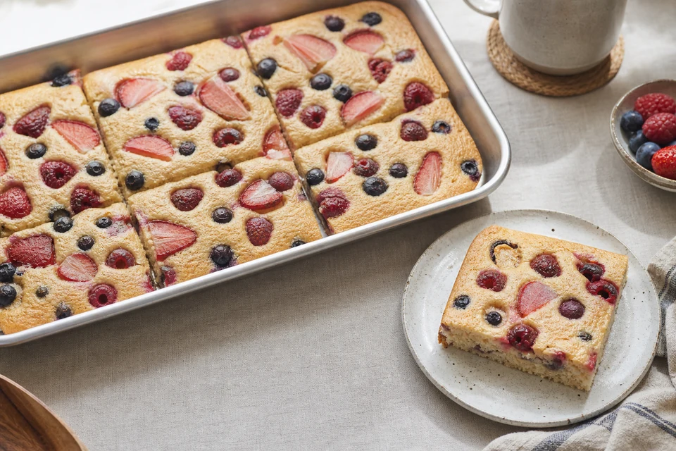 Finished vegan berry sheet-pan pancakes cut into squares with strawberries, raspberries, blueberries, and one plated piece showing the cooked crumb.