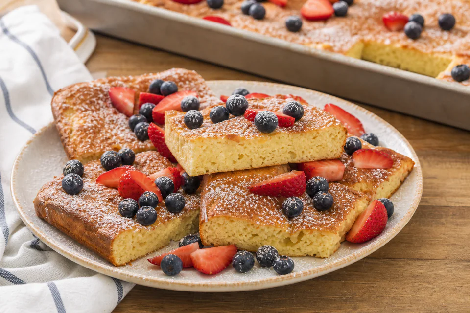Finished baked pancake squares with berries and a sheet pan visible behind the serving plate
