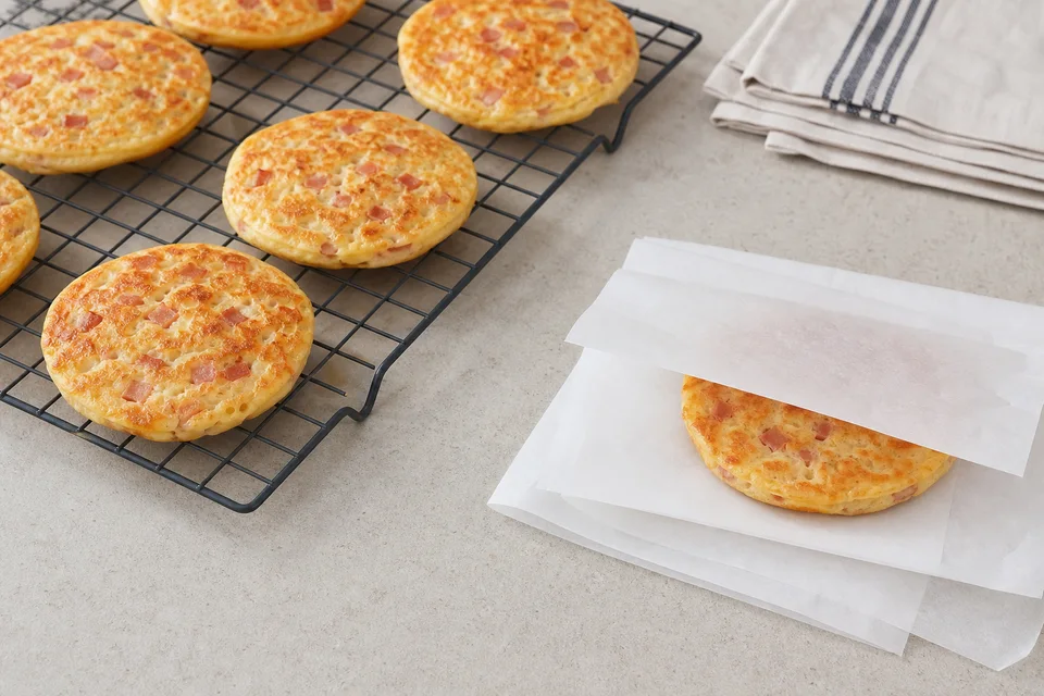 Cooked ham-and-cheese pancakes cooling on a rack beside one pancake separated with parchment for freezing.