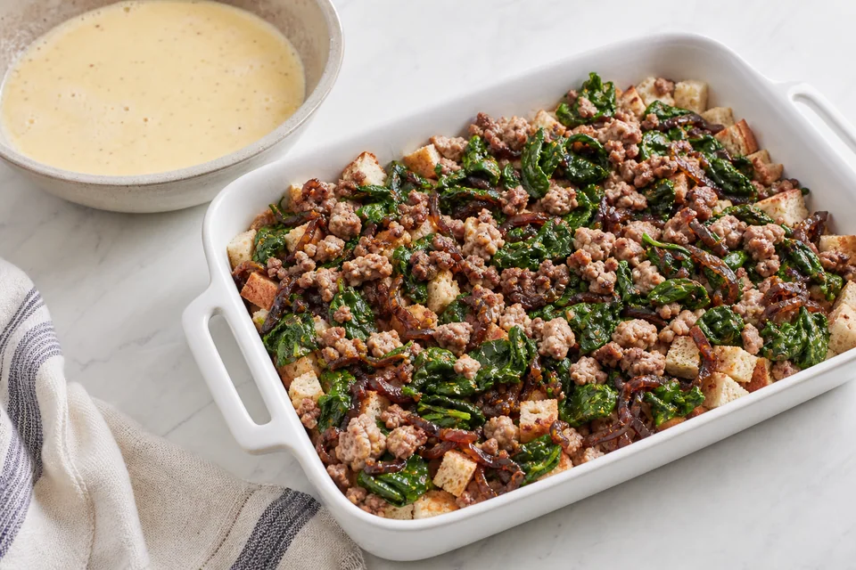 A baking dish layered with bread, browned pork, onion, and spinach beside a bowl of egg custard before baking.