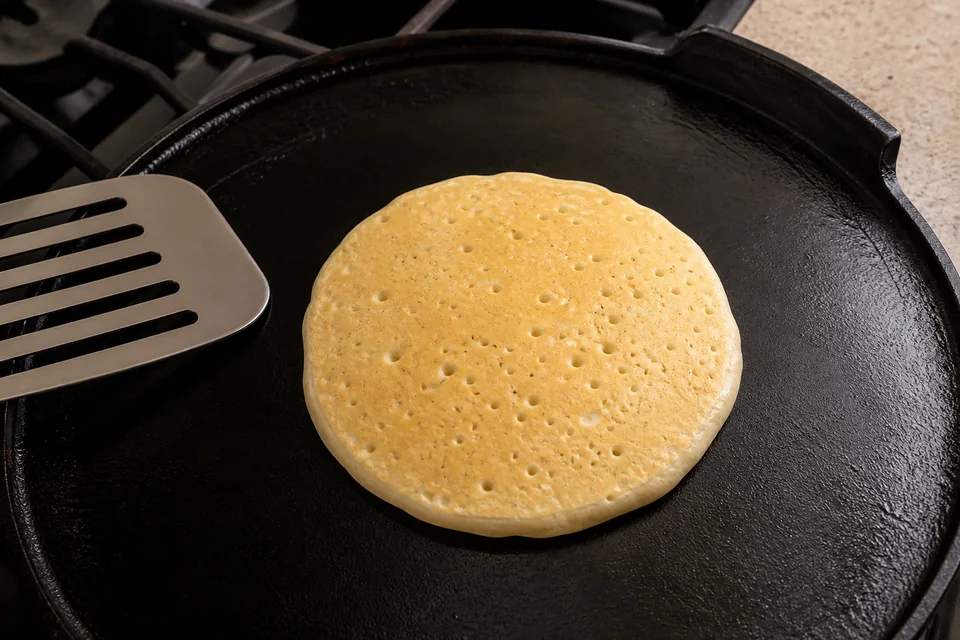 A classic American pancake on a cast-iron griddle with open bubbles and dry edges, ready to turn.