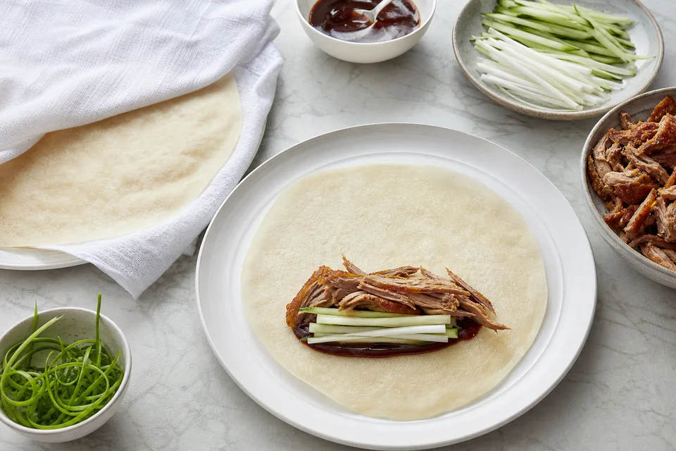 An open thin wrapper with measured sauce, cooked duck, cucumber, and scallion ready to roll.