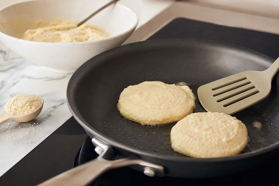 Two small almond-flour pancakes cooking in a nonstick skillet beside a bowl of thick batter and a wide spatula.