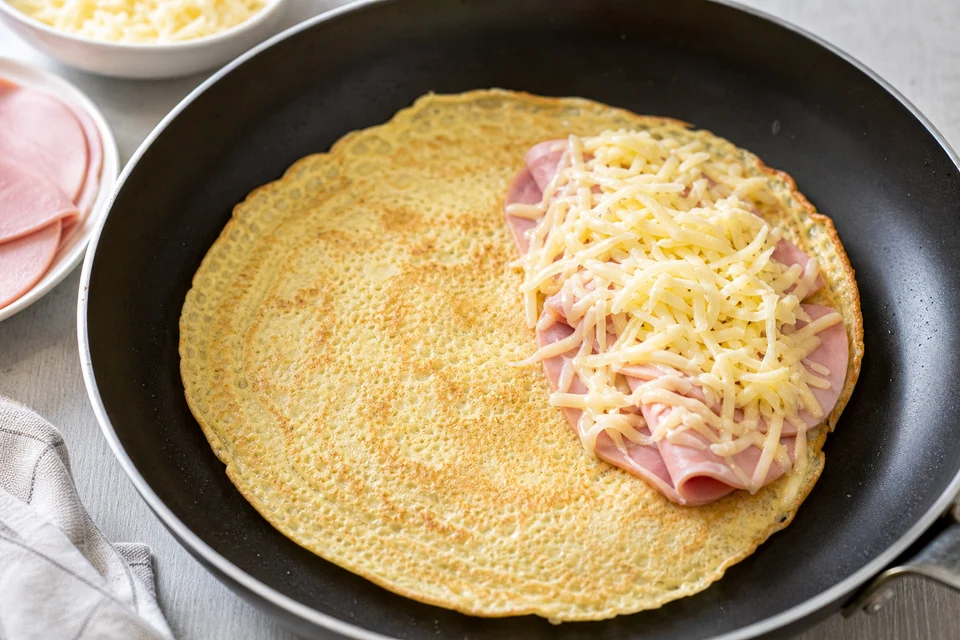 Thin savory pancake cooking with ham and grated cheese across one half.