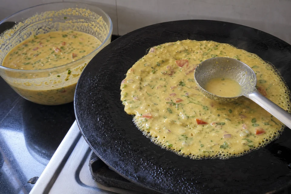 Vegetable-speckled besan chilla batter being spread thinly on a hot pan with the back of a ladle.