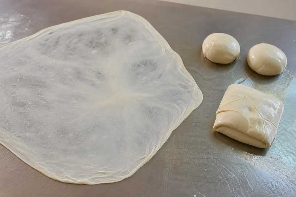 A thin sheet of roti canai dough being folded on a work surface before griddle cooking.