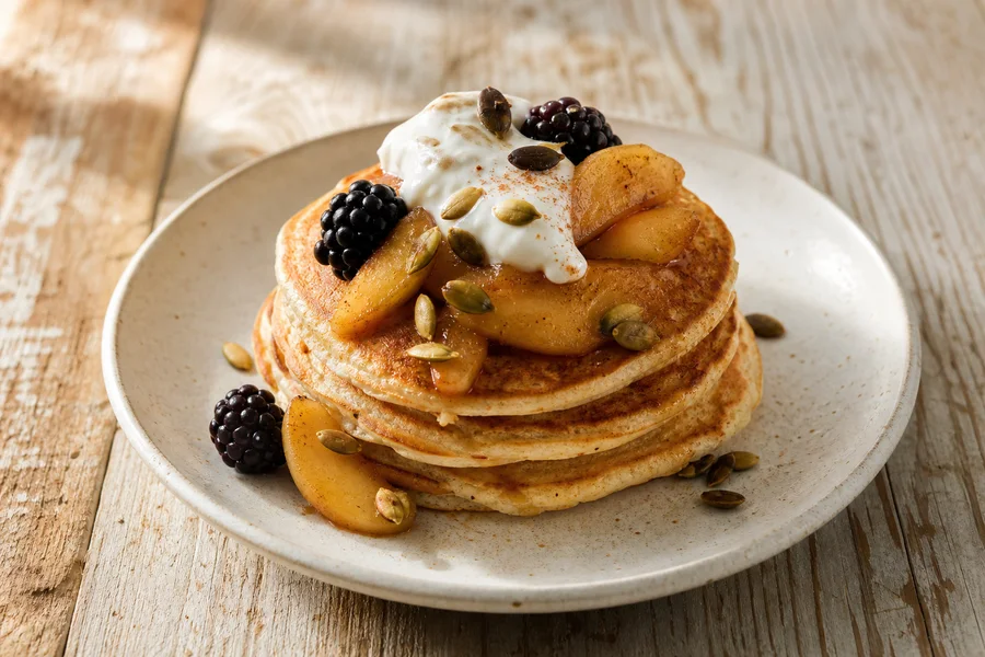 Close detail of breakfast pancakes topped with cinnamon apples, yogurt, pumpkin seeds, and blackberries.