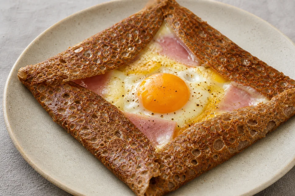 Finished buckwheat galette folded into a square around cooked ham, melted cheese, and a set egg white with crisp lacy edges.