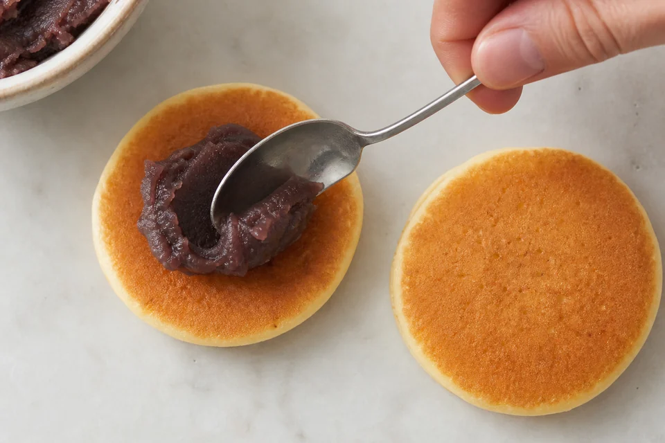 Sweet red bean paste being spooned onto a dorayaki cake before the second round is added.