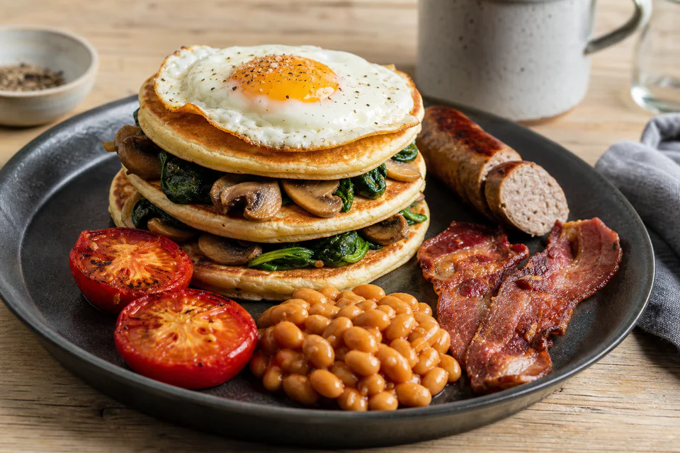 Savory pancake stack with cooked sausage, bacon, fried egg, mushrooms, tomato, baked beans, and chives