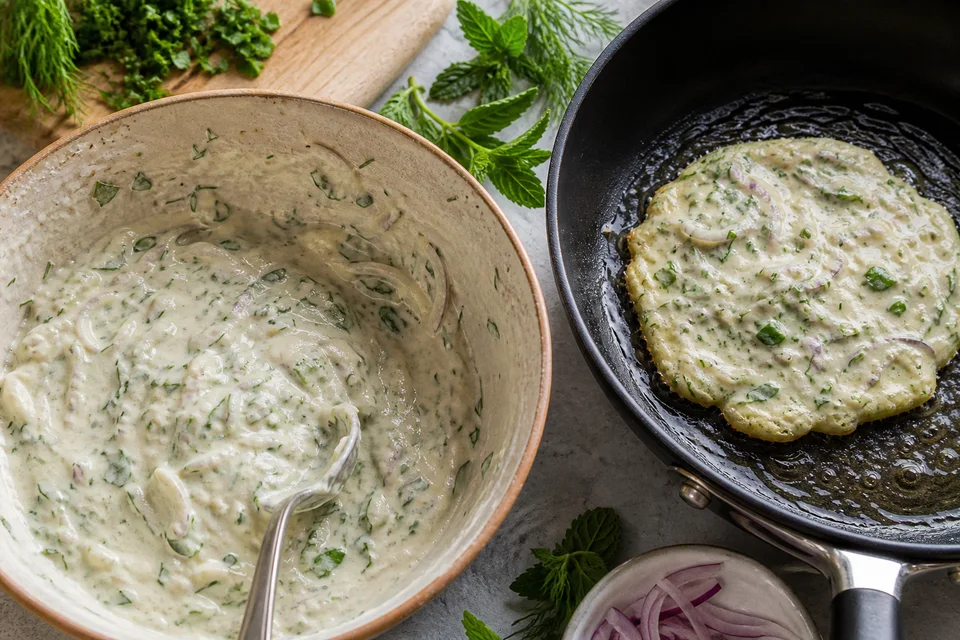 Onion-and-herb pancake batter beside one small pancake cooking until its edges set.