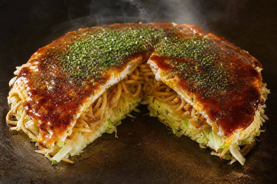 Cut Hiroshima-style okonomiyaki showing distinct cabbage, noodle, batter, and egg layers beneath glossy sauce.