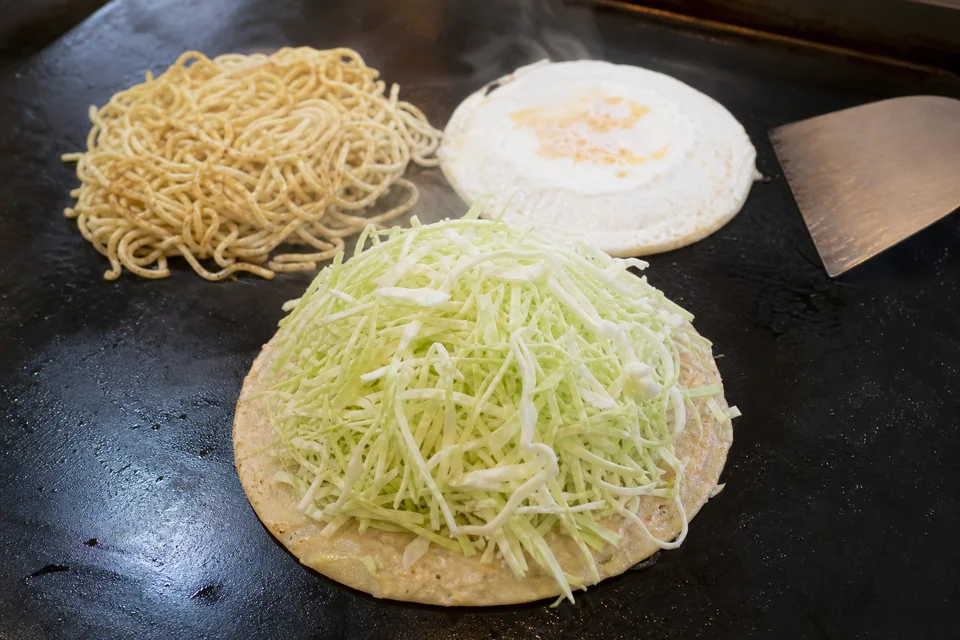 Thin batter round topped with sliced cabbage beside cooked noodles and egg during Hiroshima-style okonomiyaki assembly.