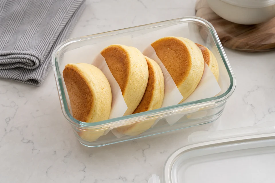 Fully cooked Japanese pancakes separated with parchment in a shallow glass container.