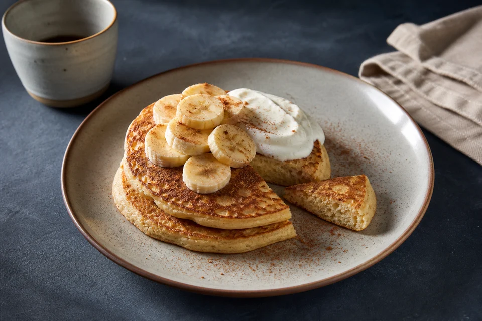 Vanilla protein pancakes with banana, yogurt, cinnamon, and a cut wedge showing the crumb.