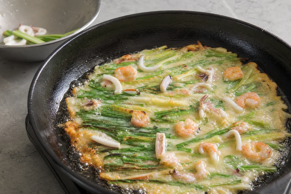 Seafood and long scallions setting in thin pajeon batter as the skillet edge bubbles and crisps.