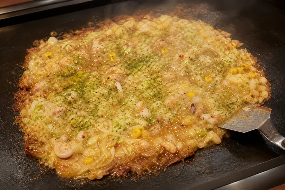 Finished monjayaki with cabbage, seafood, corn, and green seaweed seasoning cooking on a griddle.