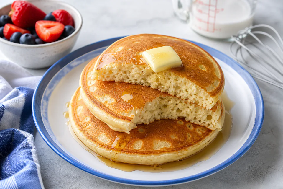 Finished fluffy pancakes cut open on a blue-rimmed plate beside berries and mixing tools