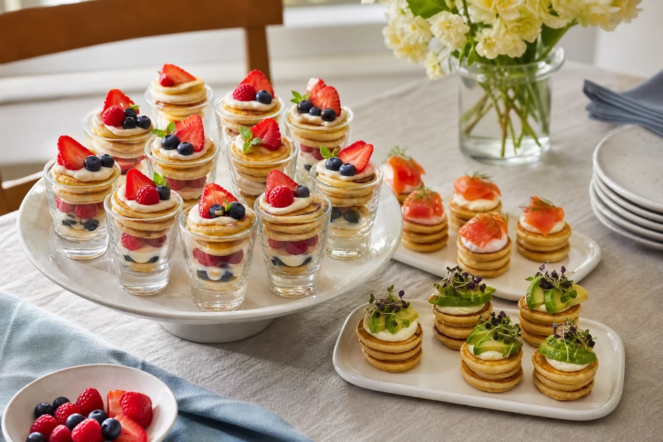 Finished bite-size pancake stacks served in small dessert glasses and on plates with berries, smoked salmon, and avocado.