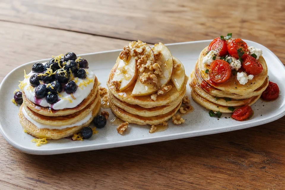 Three finished mini pancake stacks topped with blueberry lemon yogurt, pear walnut, and tomato herb cheese combinations.
