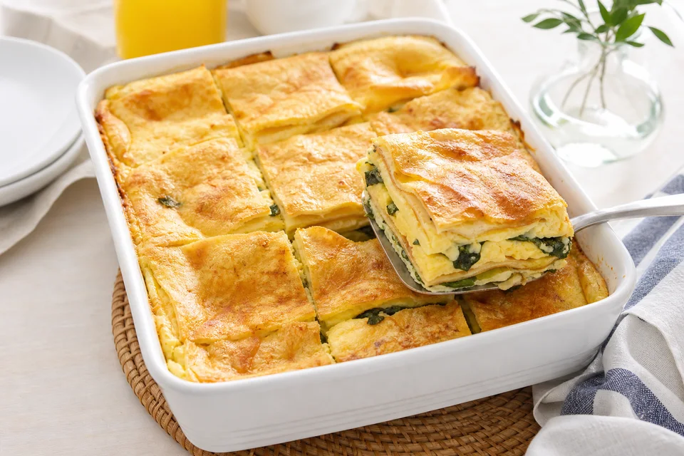 A browned scrambled-egg and crêpe breakfast casserole with one square lifted to show its layers.