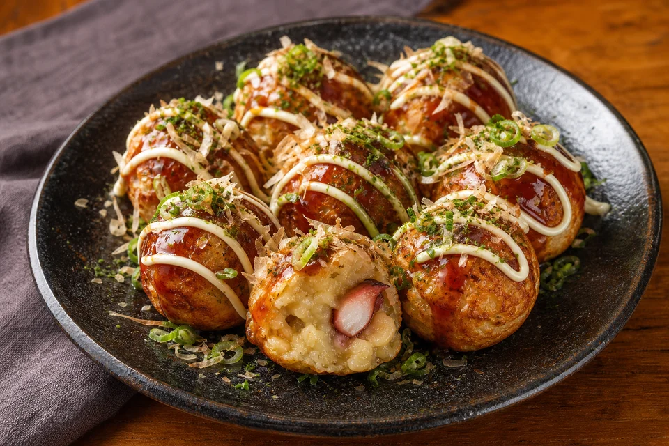 Fresh takoyaki with sauce, green onion, and bonito flakes beside a traditional dimpled cooking pan.