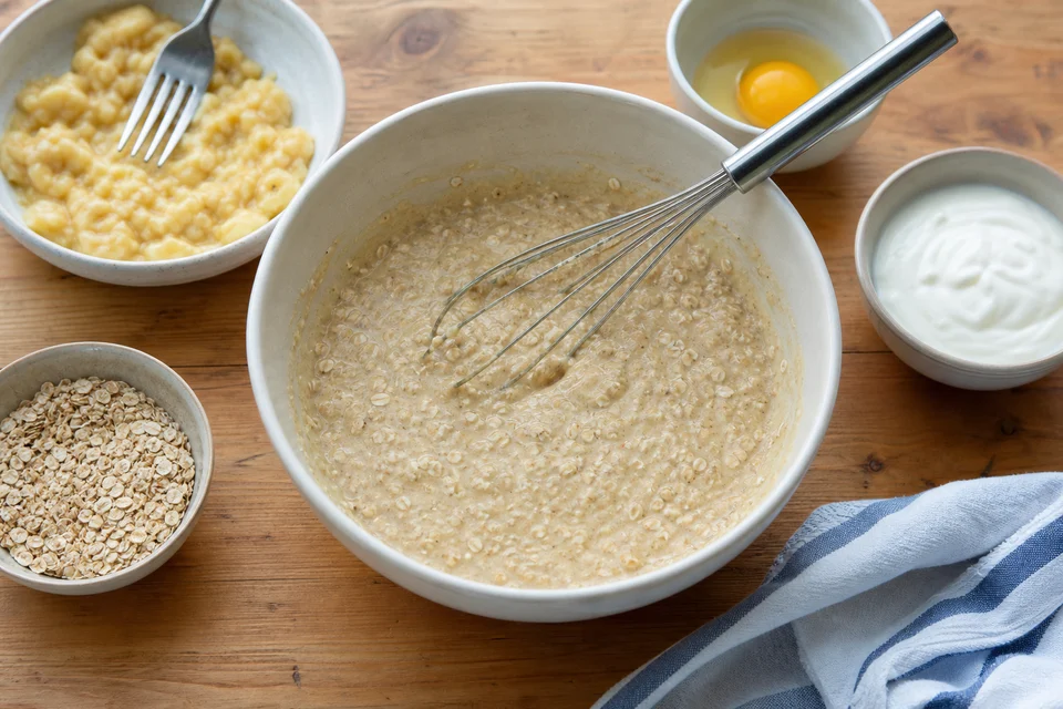 Oat flour, mashed banana, egg, yogurt, and milk arranged around a bowl of oat pancake batter.
