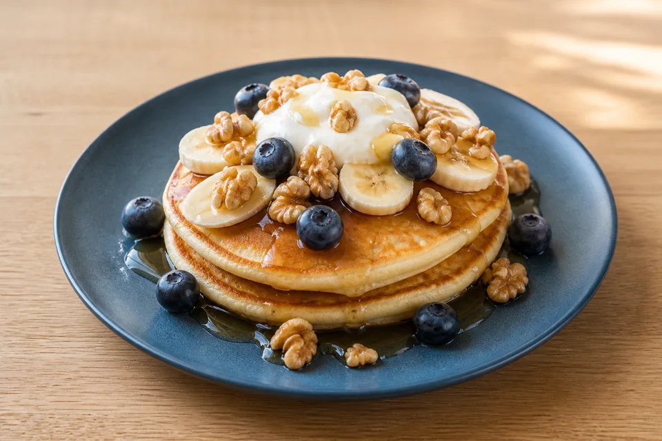 Golden pancakes topped with banana, blueberries, walnuts, yogurt, and maple syrup.