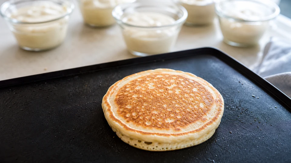 A single golden test pancake on a griddle with prepared batter portions behind it.