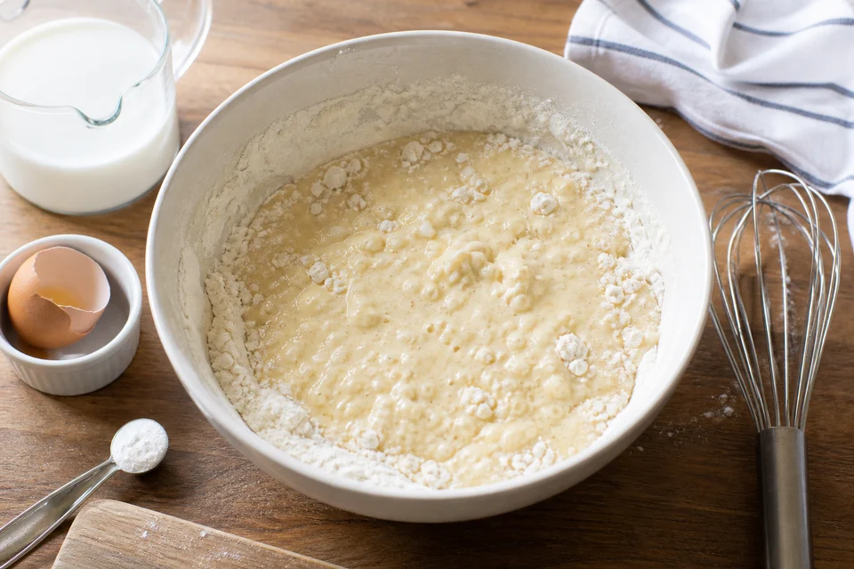 Measured flour, milk, egg, and melted butter beside a bowl of partly mixed pancake batter.