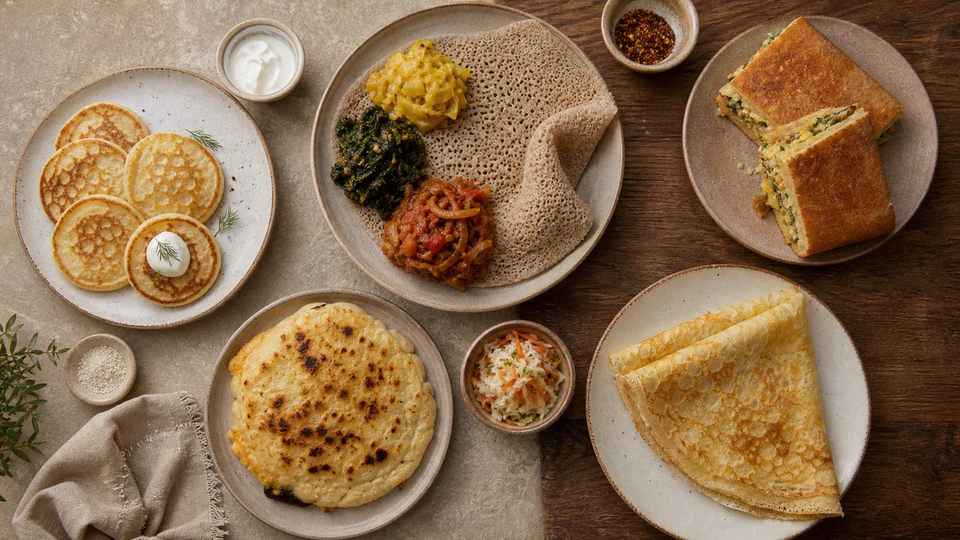 Overhead spread of pancake traditions from around the world, including injera, blini, a stuffed savory pancake, and folded crepes.
