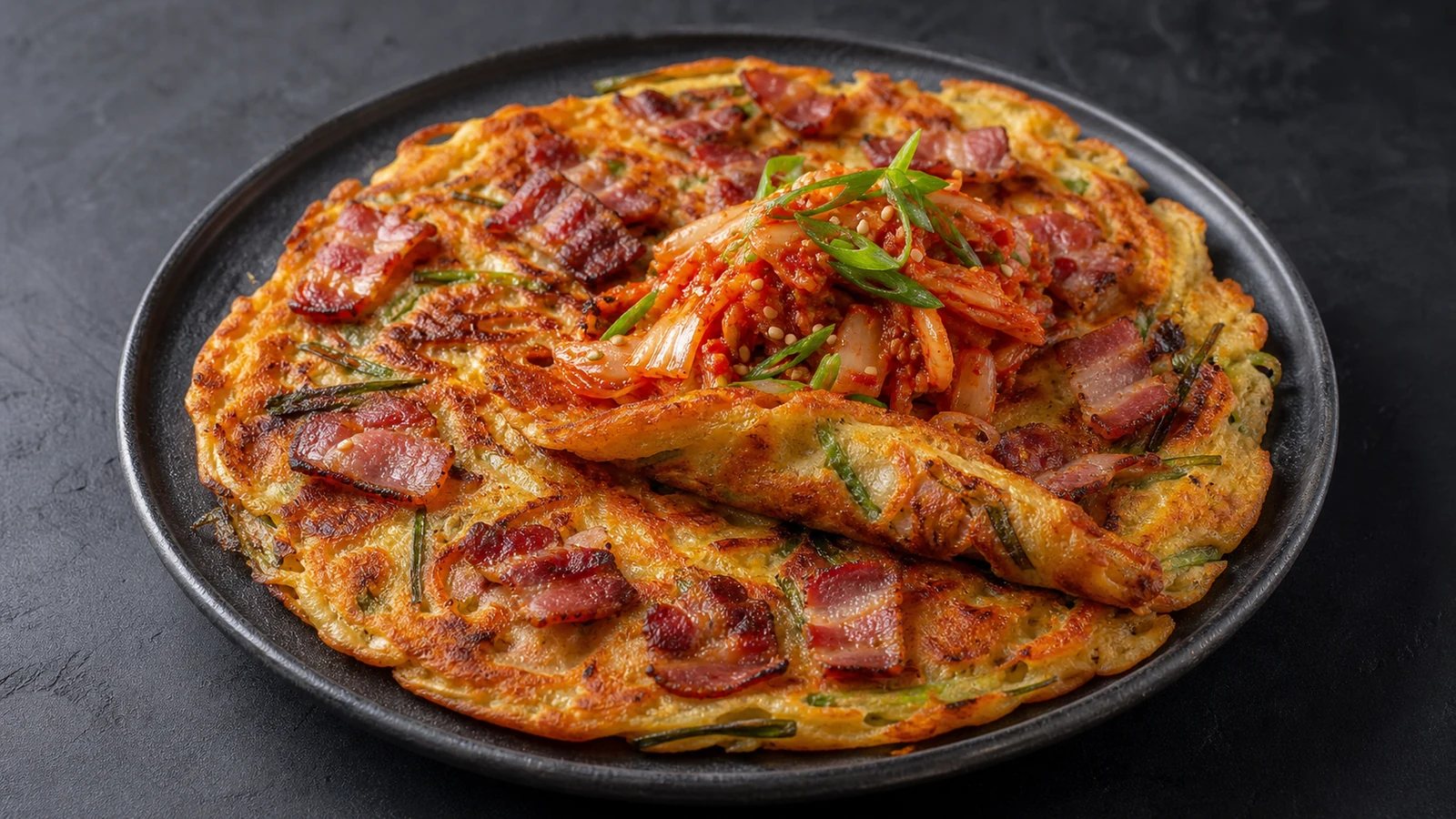 Golden savoury pancake with crisp bacon pieces, kimchi, scallions, and sesame on a dark ceramic plate.