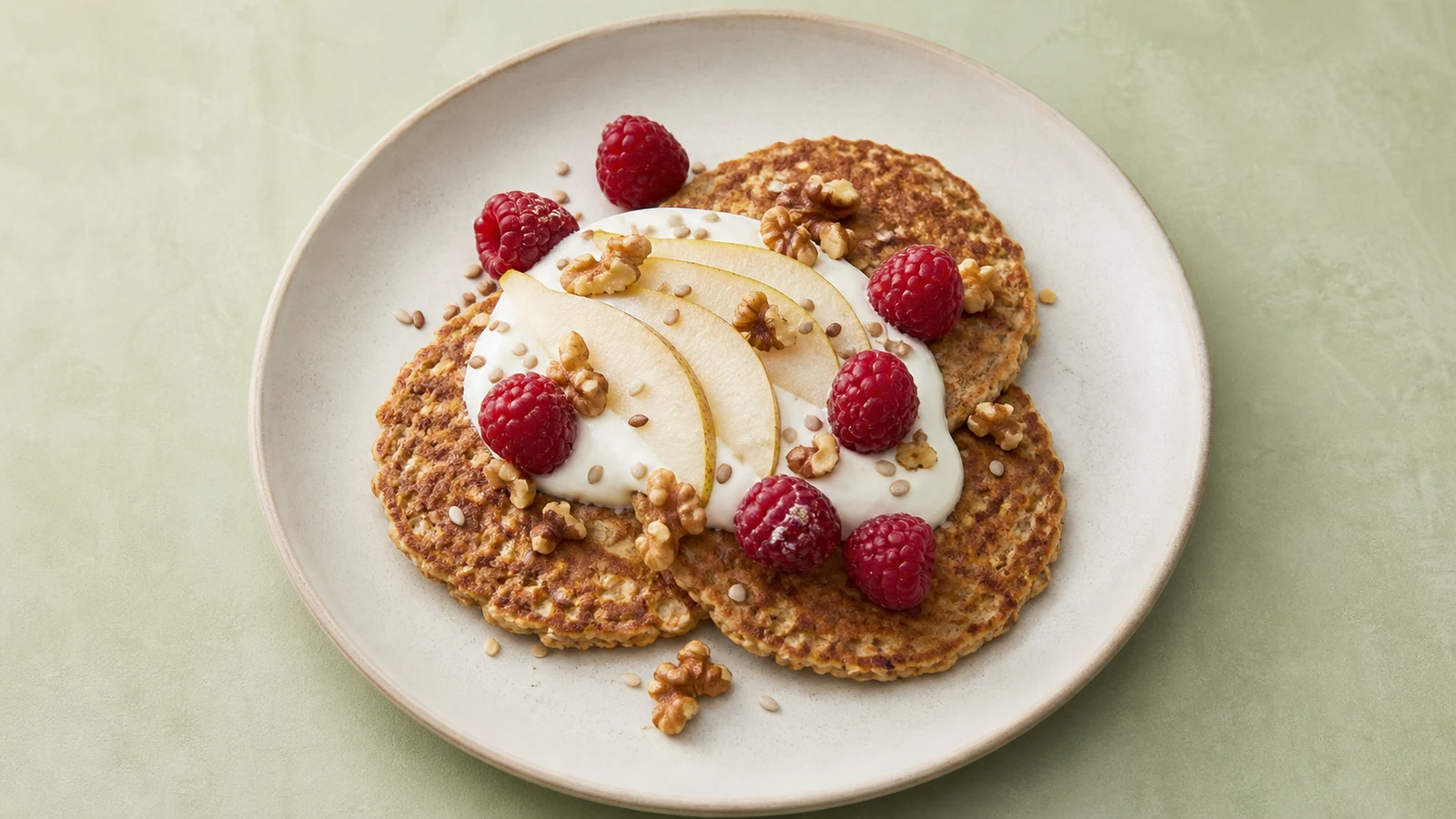 Oat-style pancakes with plain yogurt, pear slices, raspberries, walnuts, and seeds on a pale ceramic plate.