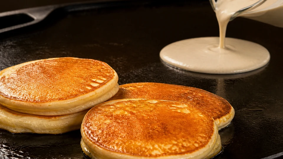 Finished pancakes beside batter pouring from a glass pitcher onto a griddle.